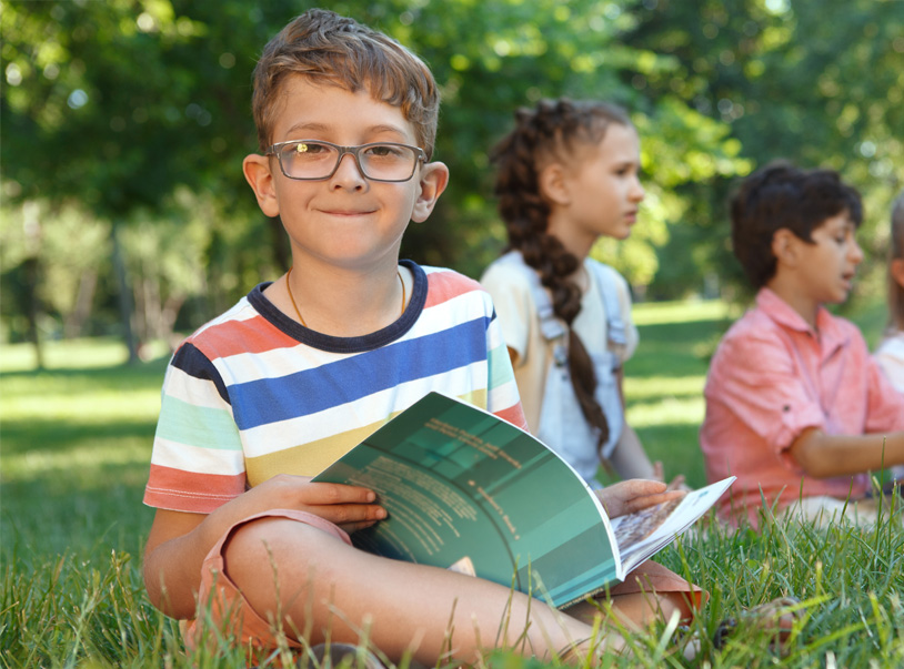 A young boy with a book smiles brightly, surrounded by friends in a sunny park